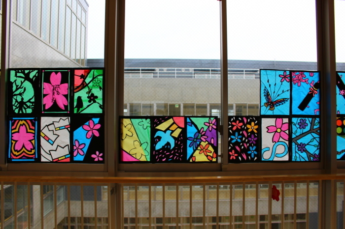 廊下の窓に、桜の花びらや、蝶、鳥などが描かれたステンドグラスの作品が飾られている写真