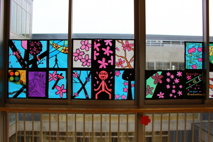 水色や黒、白の背景で、桜の木や花びら、キャラクターなどが描かれている16枚のステンドグラスの作品が窓に飾られている写真