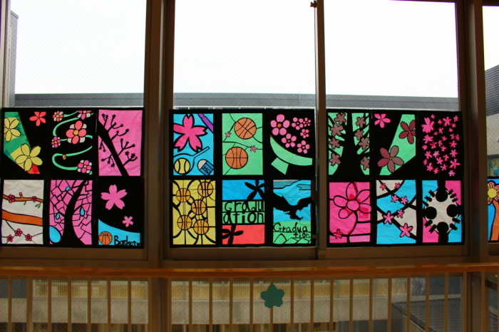 バスケットボールやラケット、花などが描かれた18枚のステンドグラスの作品が窓に飾られている写真