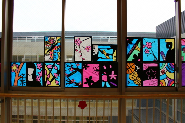 水色や白、カラフルな背景に、桜の木や、花びら、女性などが描かれている15枚のステンドグラスの作品が窓に飾られている写真