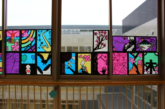 花や楽器、景色などが描かれた15枚のステンドグラスの作品が窓に飾られている写真