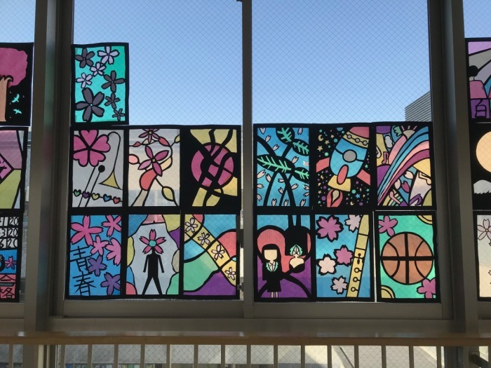 窓に桜の花びらや、ロケット、バスケットボールなどが描かれたステンドグラスの作品が飾られ、その奥に青空が写っている写真