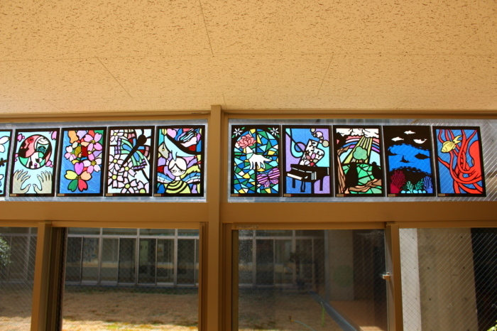廊下の上の小さい窓に、ピアノや花々、魚などが描かれたステンドグラスの作品が飾られている写真