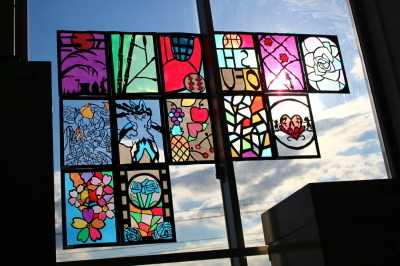 カラフルな花や、幾何学模様のステンドグラスの作品が飾られた窓の隙間から青空が見えている写真