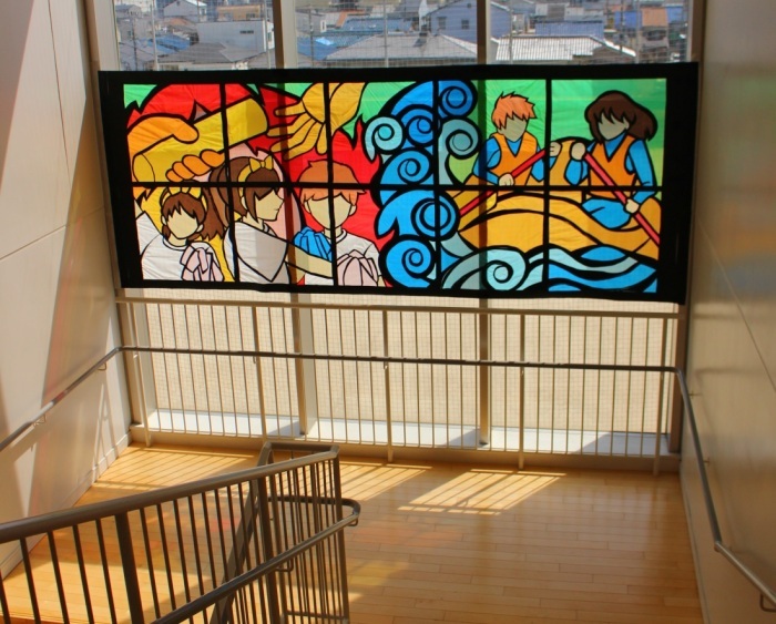 階段の踊り場の大きな窓に、バトンを渡す手や、応援団、高波でボートをこぐ様子が描かれた大きなステンドグラスの作品が飾られいる写真