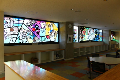 体育祭の様子や、学生生活を描いた大きなステンドグラスに作品2枚と、その間に小さな作品が飾られている室内の写真