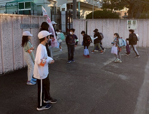 校門の外側に白色の帽子を被った児童が立ち登校してくる児童たちにあいさつ運動を行っている様子の写真