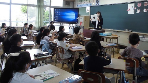 黒板の前で女性教師が中央に黒丸が描かれた白色の用紙を持っているのを児童たちが見ている授業参観の様子の写真