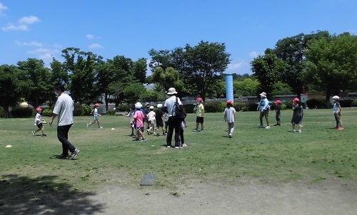 木々に囲まれた芝生広場で児童たちが走り回って遊んでいる様子の写真