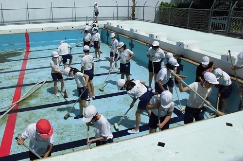 赤白帽子を被った6年生がデッキブラシを使ってプールの掃除を行っている様子の写真