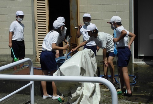 児童がホースから出てくる水をバケツに貯めているのを5名のクラスメイトが見ている写真