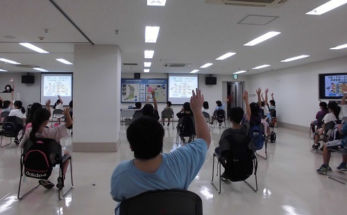 スクリーンが設置された部屋に間隔をあけて座っている児童たちが手を挙げている後ろ姿の写真