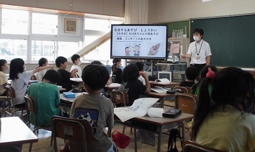 道徳の授業参観で黒板の左側に設置された大型テレビに映し出された資料を見ながら男性教師の話を席に座って聞いている2年生の児童の写真