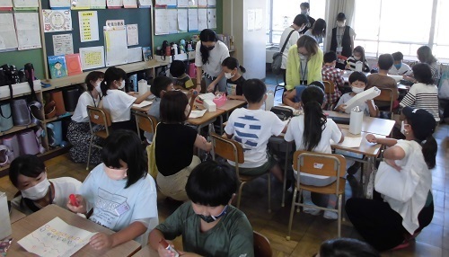 教室でグループごとに分かれて座っている3年生の児童が保護者と一緒に紙パックに絵を書いている工作の授業参観の様子の写真