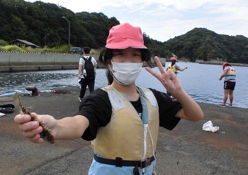 赤白帽子を被った女児が自分で釣った魚を右手に持ってピースサインをしている写真