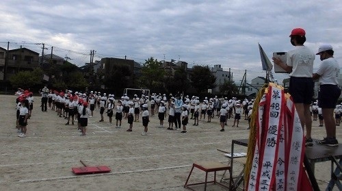 運動場に赤白帽子に体操着を着た児童たちが列を作って立ち開会式の練習を行っている様子の写真