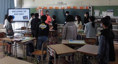 他の児童が見ているなか教室の黒板の前に立っている男性教師と男の子が右手を挙げているオープンスクールの様子の写真