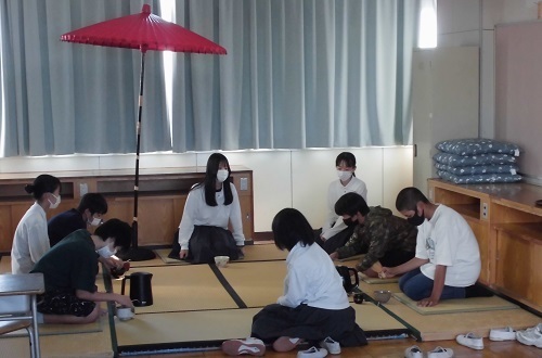 赤色の和傘が置いてある畳敷きの部屋で4名の男の子が女子生徒が見ているなかお茶をたてている茶道部の体験の様子の写真
