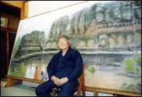 大きな絵画の作品の前の椅子にすわっている直原玉青画伯の写真