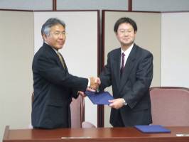 左に藤川教育長、右に谷本学長が握手を交わし協定書を持って立っている様子を写した写真