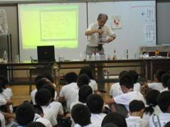 横浜国立大学の廣田名誉教授が、下島小学校の児童の前で野菜や果物のしぼり汁を使った実験を行っている様子を写した写真