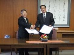 藤川博史教育長と長尾彰夫学長が立って握手をしている様子を写した写真
