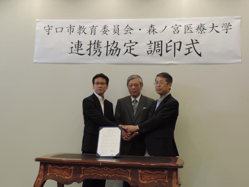 協定書を前に、中央に首藤教育長、左に清水理事長、右に萩原学長が立っている様子を写した写真