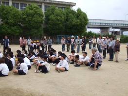 下島小学校の6年生児童が校庭に集まり、土の上に座りながら話を聞いている姿と、それを取り囲みながら見守る大人たちの写真