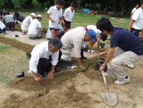 レンガの小道付近にある掘り起こされた土の部分にポット苗を植えている作業員や大人たちの写真