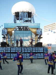 大きなステージが設置されている前で青い法被を着た人たちが提灯踊りを踊っている写真