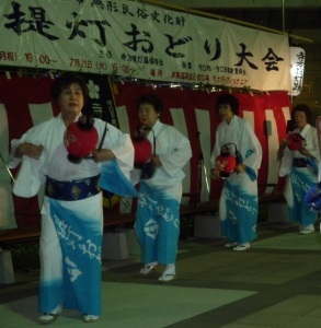 「提灯おどり大会」と書かれた幕の下で、白と水色の浴衣を着て赤い提灯を持った女性の方々が踊りを踊っている写真