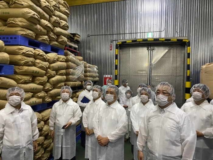 視察の参加者の方々が米袋が高く積まれた工場内の見学をしている様子の写真