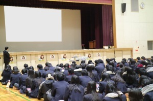 生徒たちが体育館に集まり、並んで座り講義を受けている様子を後方より写した写真