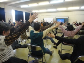 地域包括支援センターの室内で椅子に座り、両手を広げ体操をする数十名の高齢者の方々の写真