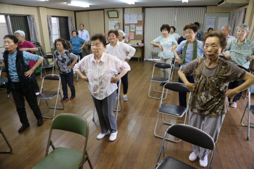 集会所で多くの高齢者が腰に手をあてて立っている写真