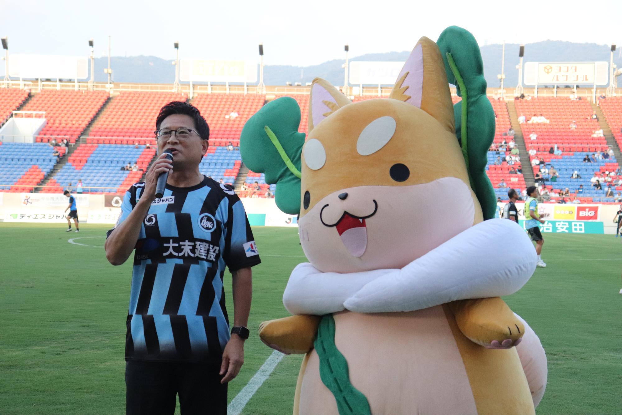 グラウンドでスピーチする市長とその横にもり吉