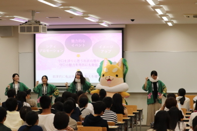 関西大学オープンキャンパスでスクリーンの前に立つもり吉と4名の法被を着た女性の写真