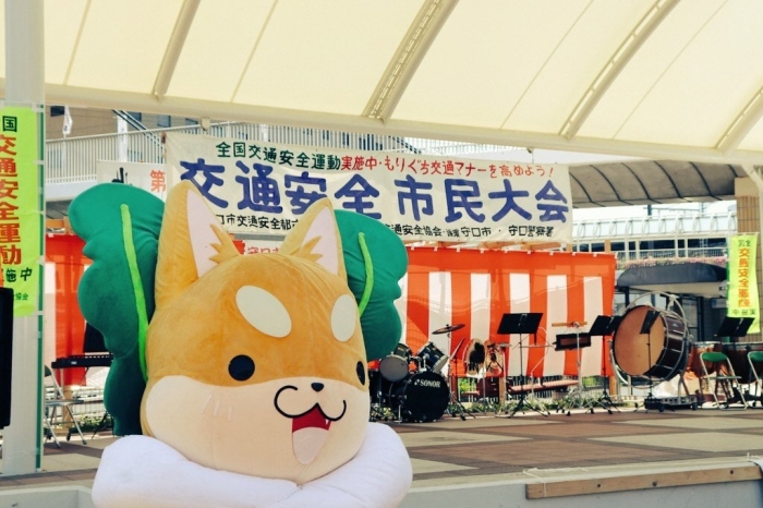 交通安全市民大会と書かれた幕の前にたくさんの楽器が並んでいる舞台近くにもり吉が立っている写真