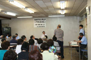 参加者の中の1人の男性が立ちながら質問をしているタウンミーティングの様子を写した写真